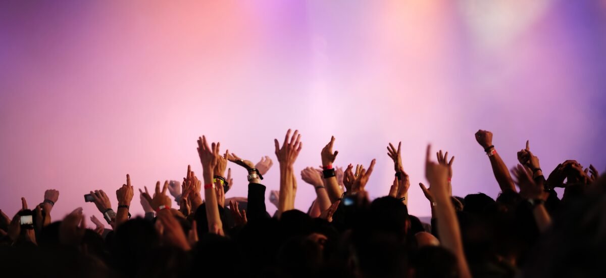 Crowd with raised hands at a concert, colorful lighting in background.