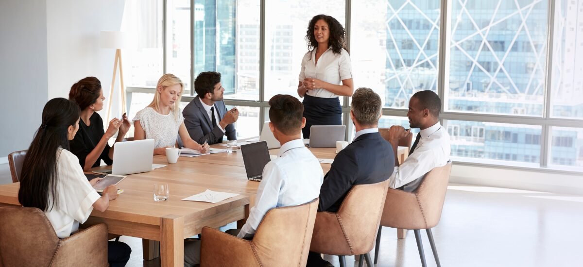 Woman speaking to colleagues at a conference table with laptops in a modern office setting.