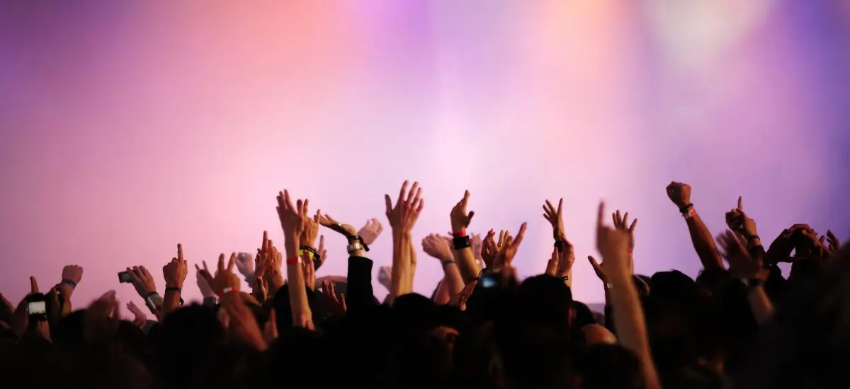 Crowd with raised hands at a concert, colorful lighting in background.